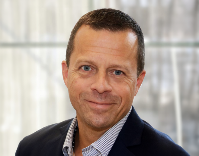 Man i ljus skjorta och mörk kostym som ler mot kameran. Ljus bakgrund.