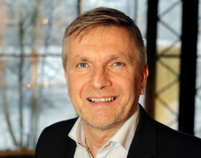 Man i ljus skjorta och svart kavaj som ler mot kameran.