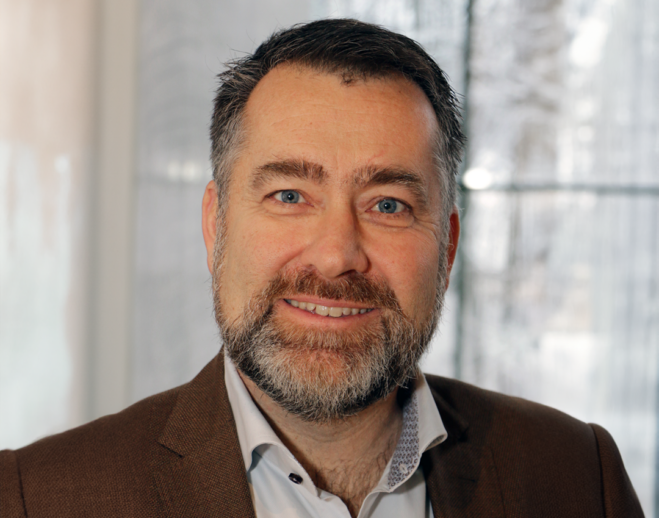 Man i ljus skjorta och mörk kostym som ler mot kameran. Ljus bakgrund.