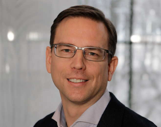 Man i jus skjorta, mörk kostym och glasögon som ler mot kameran. Grå bakrgund.