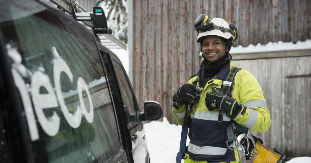 En OneCo-tekniker som ler mot kameran när han står bredvid en öppen bildörr. Teknikern har på sig vit hjälm och säkerhetsutrustning.