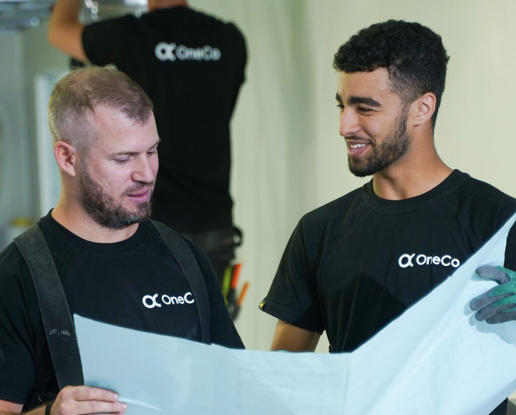 Två OneCo-tekniker i svara t-shirtar som håller i en karta tillsammans. Den ena ler och den andra pratar. I bakgrunden syns ytterligare en tekniker som har ryggen vänd mot kameran.