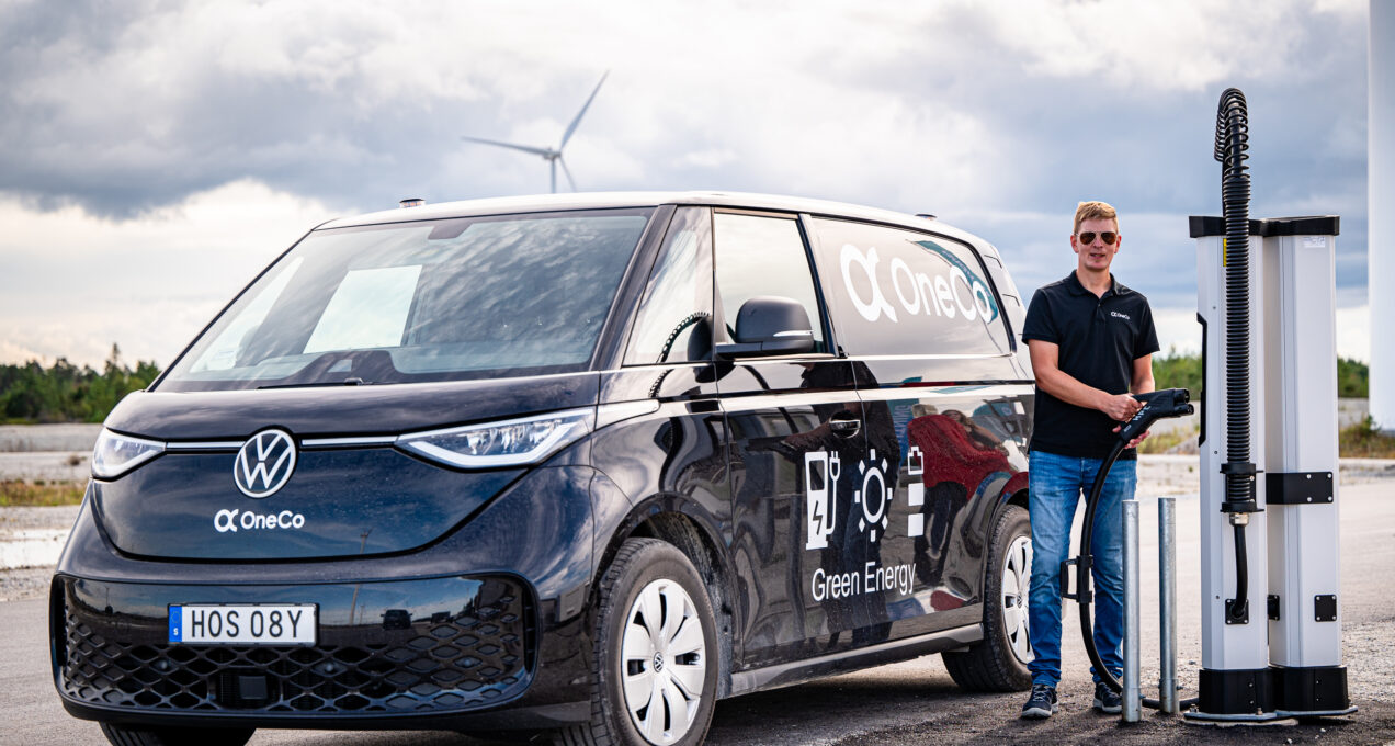 En man som står och håller i en DC-laddare. Bredvid mannen står en svart OneCo-bil.