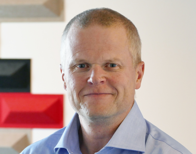 Man i ljusblå skjorta som ler mot kameran. Vit, röd och svart bakgrund.