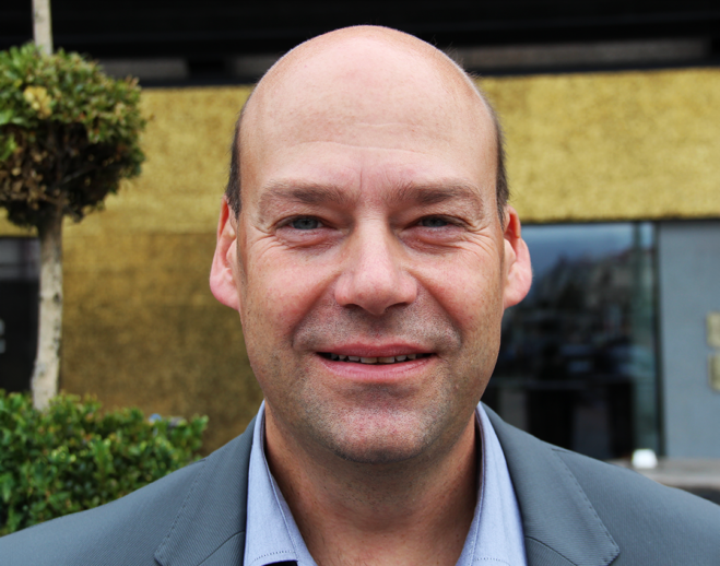 Man i grå kavaj och blå skjorta som ler mot kameran. I bakgrunden skymtar ett hus.