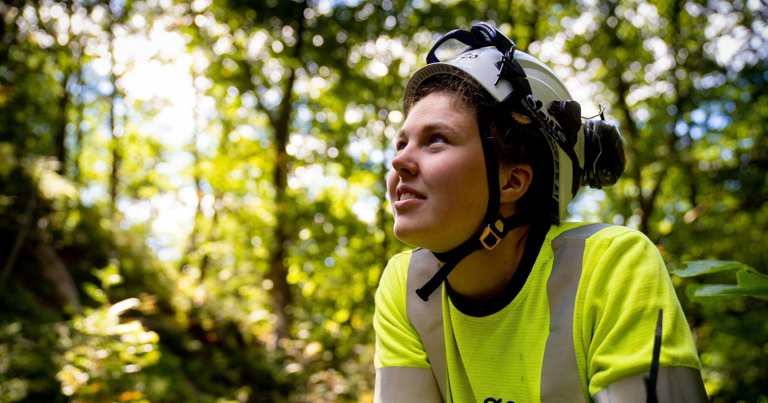 OneCo-tekniker i gul tröja och vit hjälm i en skog som tittar snett uppåt.
