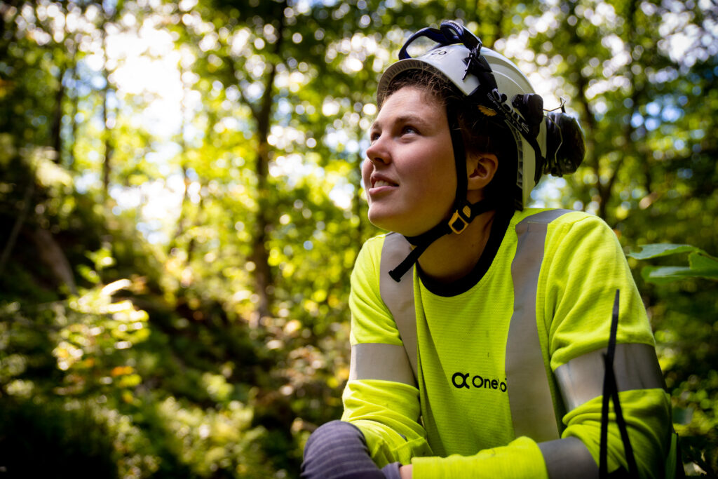 OneCo-tekniker i gul tröja och vit hjälm i en skog som tittar snett uppåt.