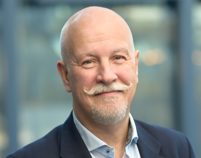 Man i blå skjorta och mörk kavar som ler mot kameran. blå, grå, grön bakrgund.