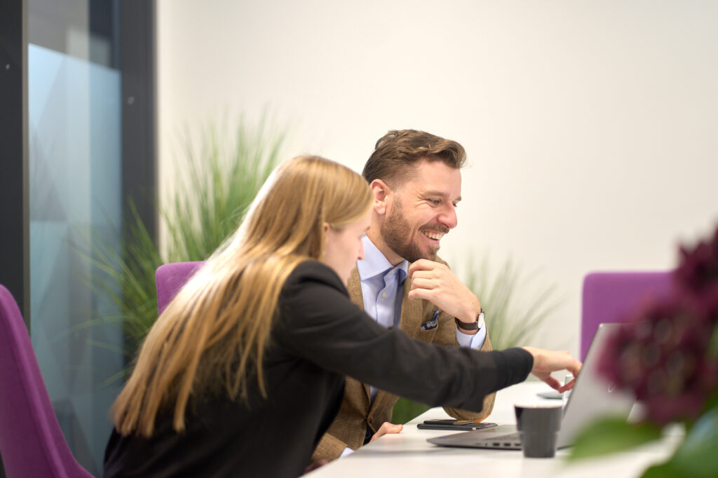 En kvinna och en man i kontorsmiljö. Kvinnan visar mannen något på et datorskärm och mannen skrattar.