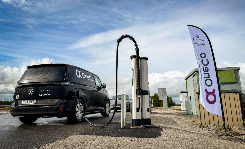 En svart OneCo-bil bakifrån som laddas med en DC-laddare. Framför OneCos bil syns en grå bil. Bredvid laddaren syns en OneCo-flagga. I bakgrunden syns blå himmel med moln.