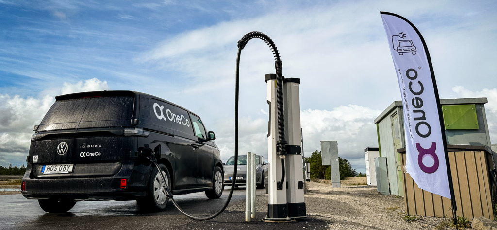 En svart OneCo-bil bakifrån som laddas med en DC-laddare. Framför OneCos bil syns en grå bil. Bredvid laddaren syns en OneCo-flagga. I bakgrunden syns blå himmel med moln.