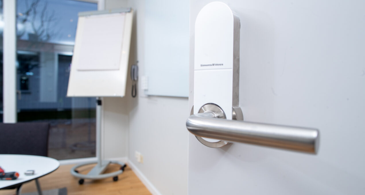 Vit dörr med digitalt lås. I bakgrunden syns ett konferensrum med ett blädderblock på ett ställ.