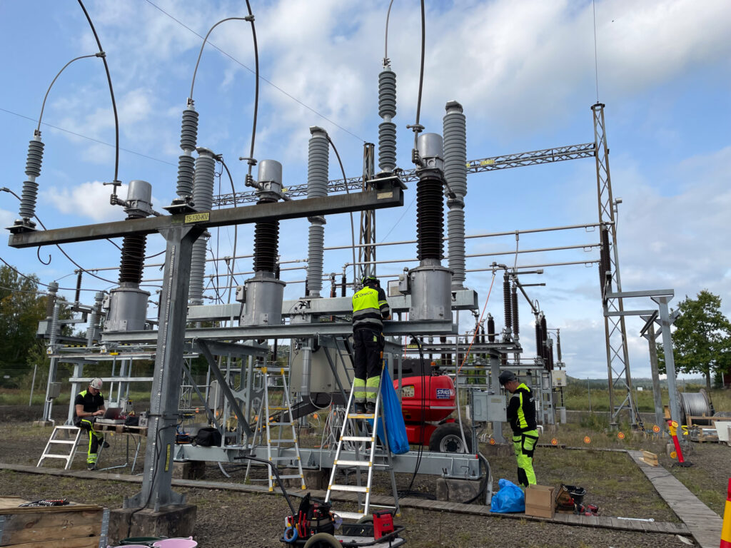 Tre OneCo-tekniker som arbetar i olika delar av ett ställverk. I bakgrunden syns blå himmel med moln.