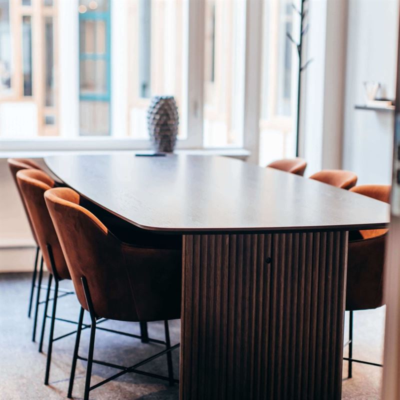 Ett ljust rum med ett stort, mörkt bord i mitten. Runt bordet står orangea stolar.