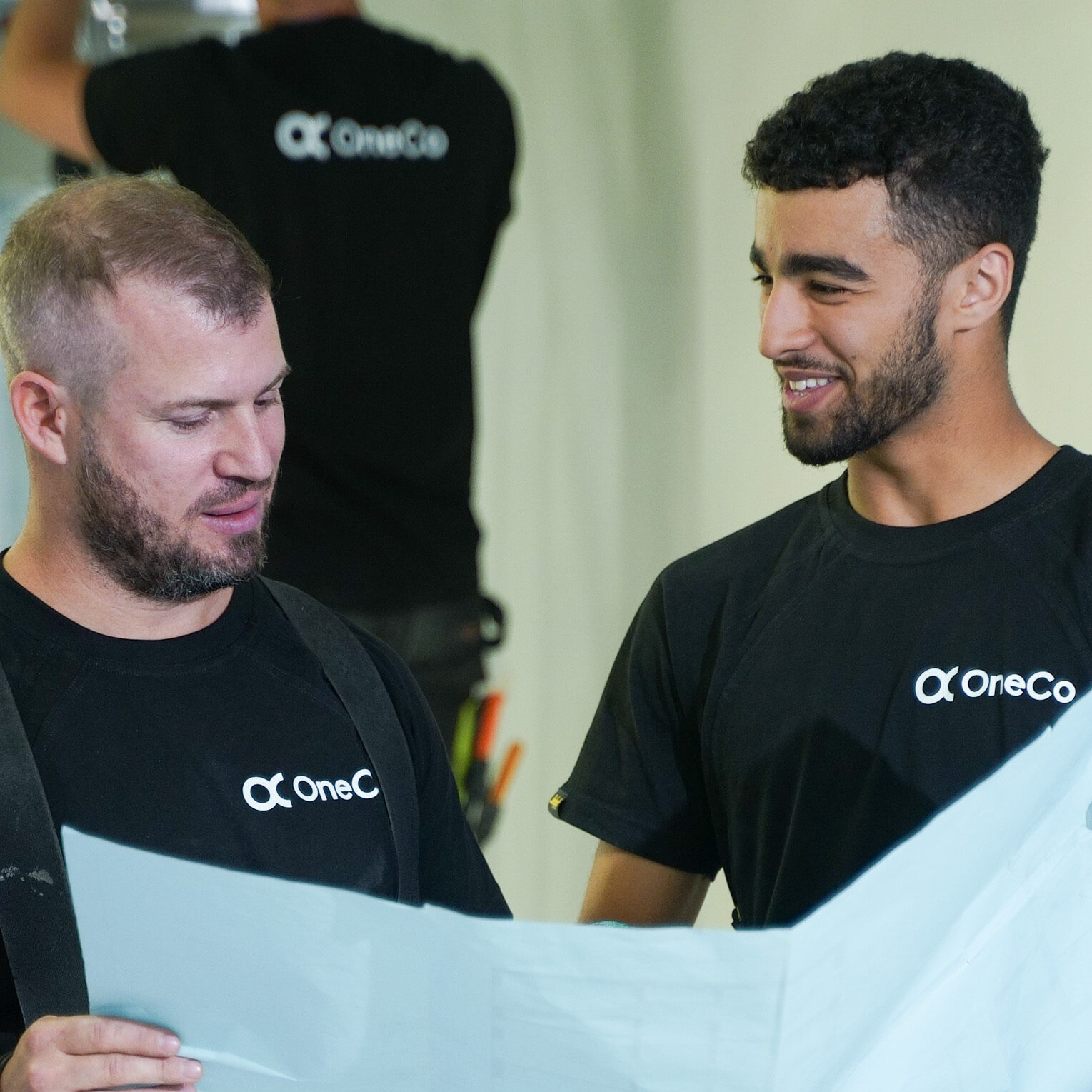 Två OneCo-tekniker i svara t-shirtar som håller i en karta tillsammans. Den ena ler och den andra pratar. I bakgrunden syns ytterligare en tekniker som har ryggen vänd mot kameran.